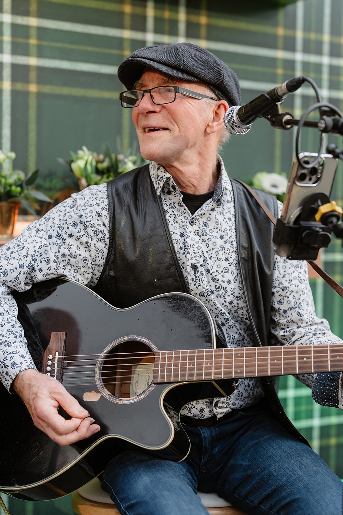 Older man in glasses and a cap plays a black acoustic guitar and smiles while sitting in front of a microphone, with a green plaid background and flowers behind him, evoking the warm, inviting atmosphere one might find at an Irish castle wedding at Ashford Castle.