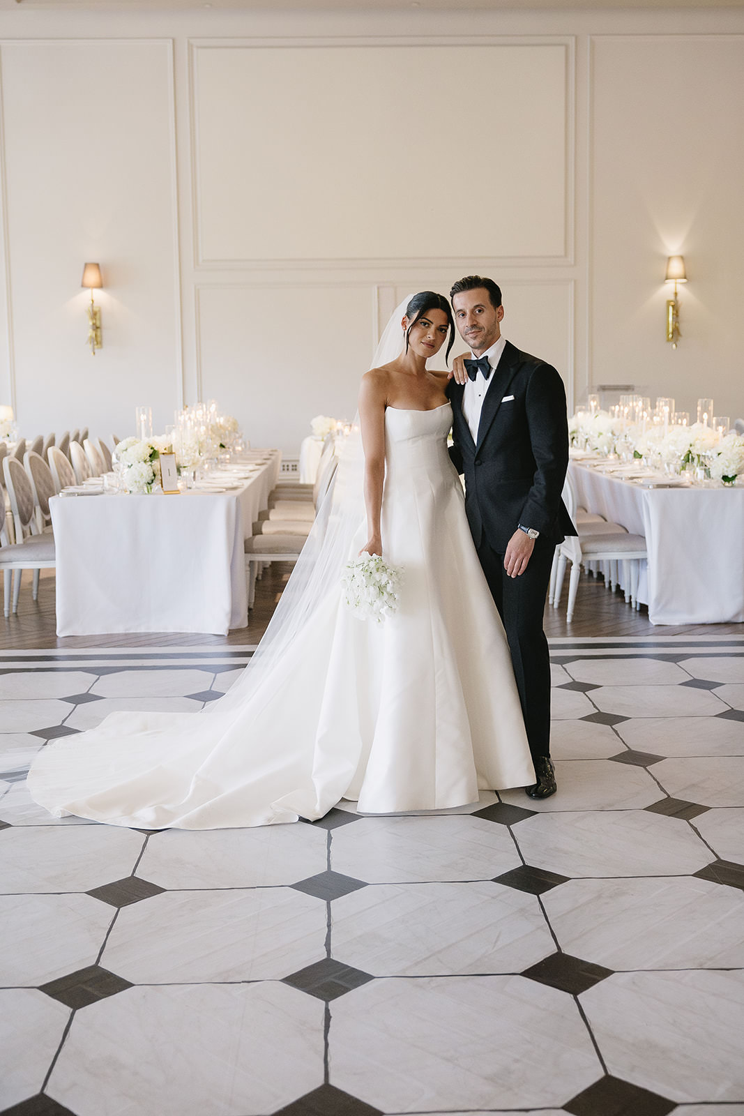 A bride in a white gown and a groom in a black tuxedo stand together in an elegant, candlelit reception hall with white decor and floral arrangements, radiating joy as they celebrate their love at Erica’s unforgettable wedding.