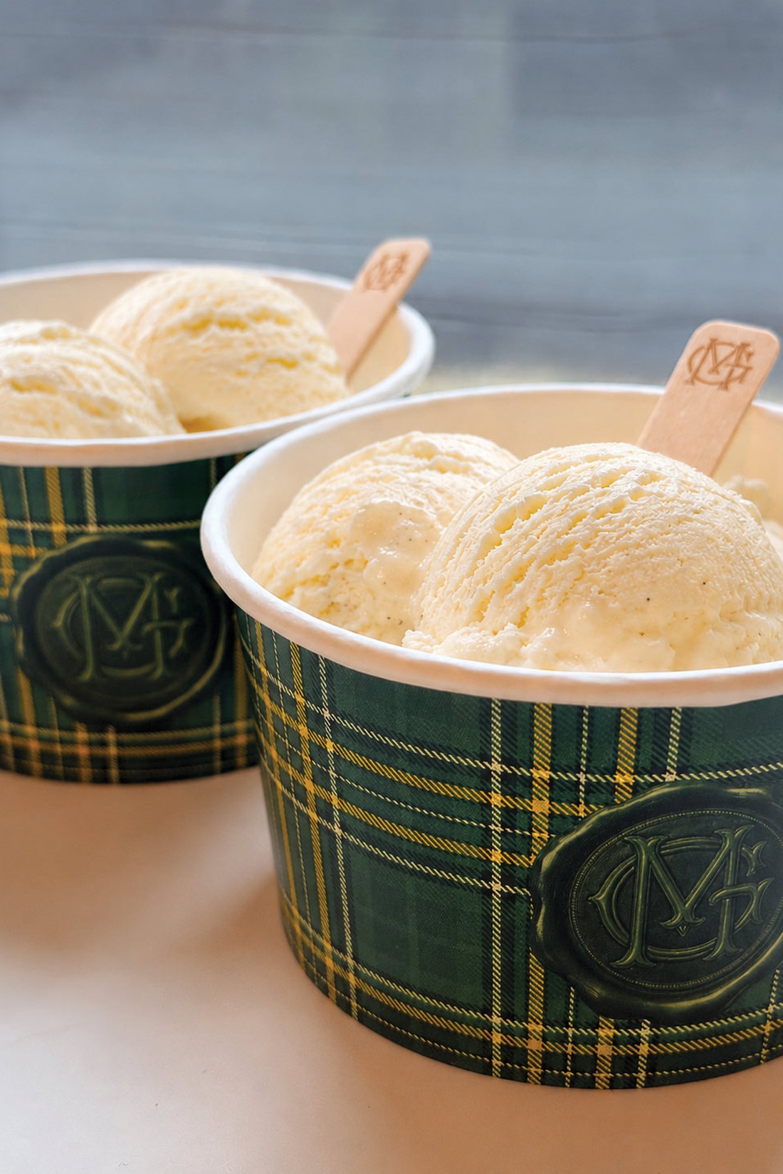 Two cups with green and yellow plaid pattern hold scoops of vanilla ice cream, each with a wooden spoon inside. The cups display a circular monogram logo on the side, evoking the charming details you might find at an Irish castle wedding at Ashford Castle.
