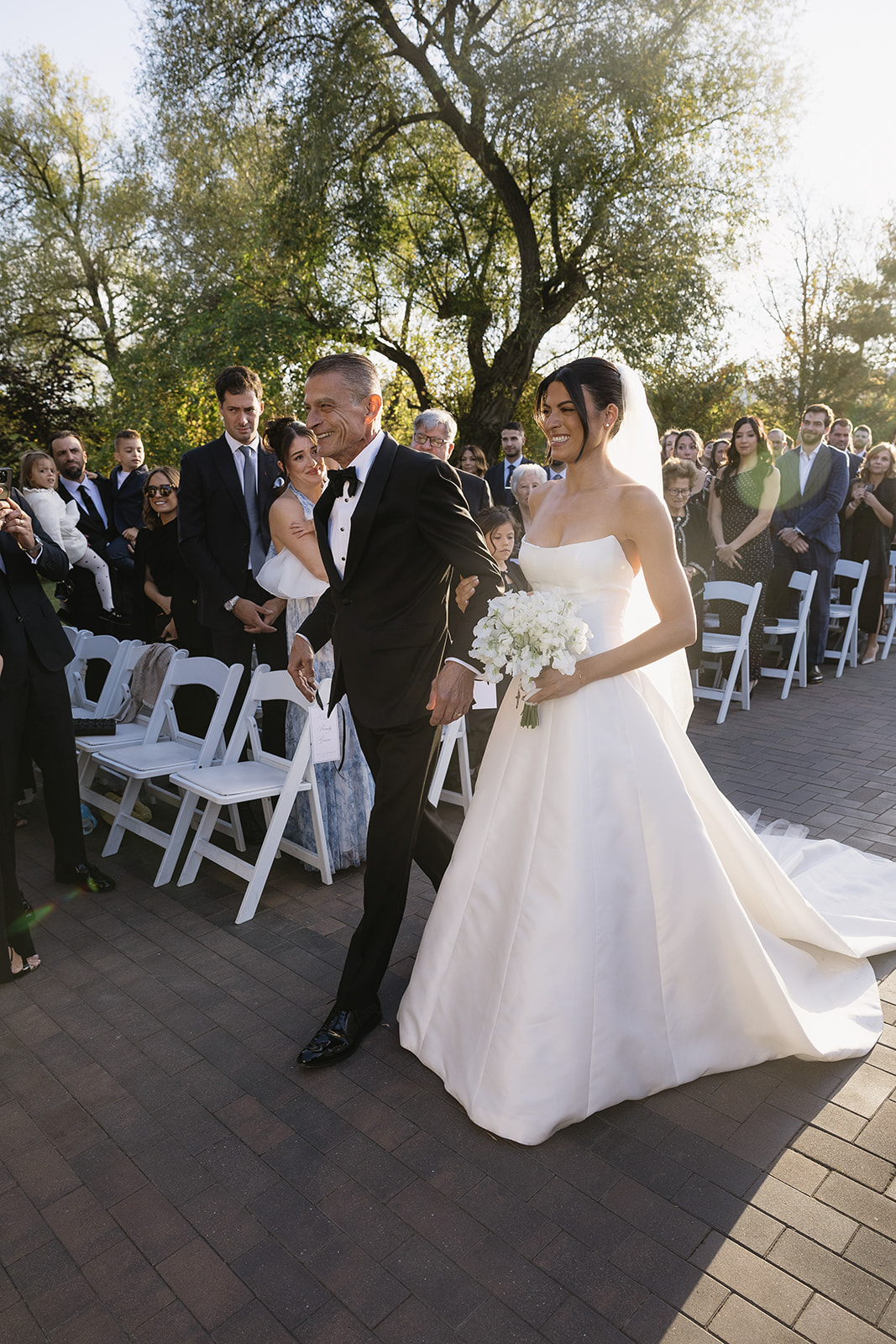 A bride named Erica, in a white gown and veil, walks down the aisle outdoors, smiling and holding a bouquet as she is escorted by an older man in a tuxedo. Guests stand and watch as sunlight filters through the trees.