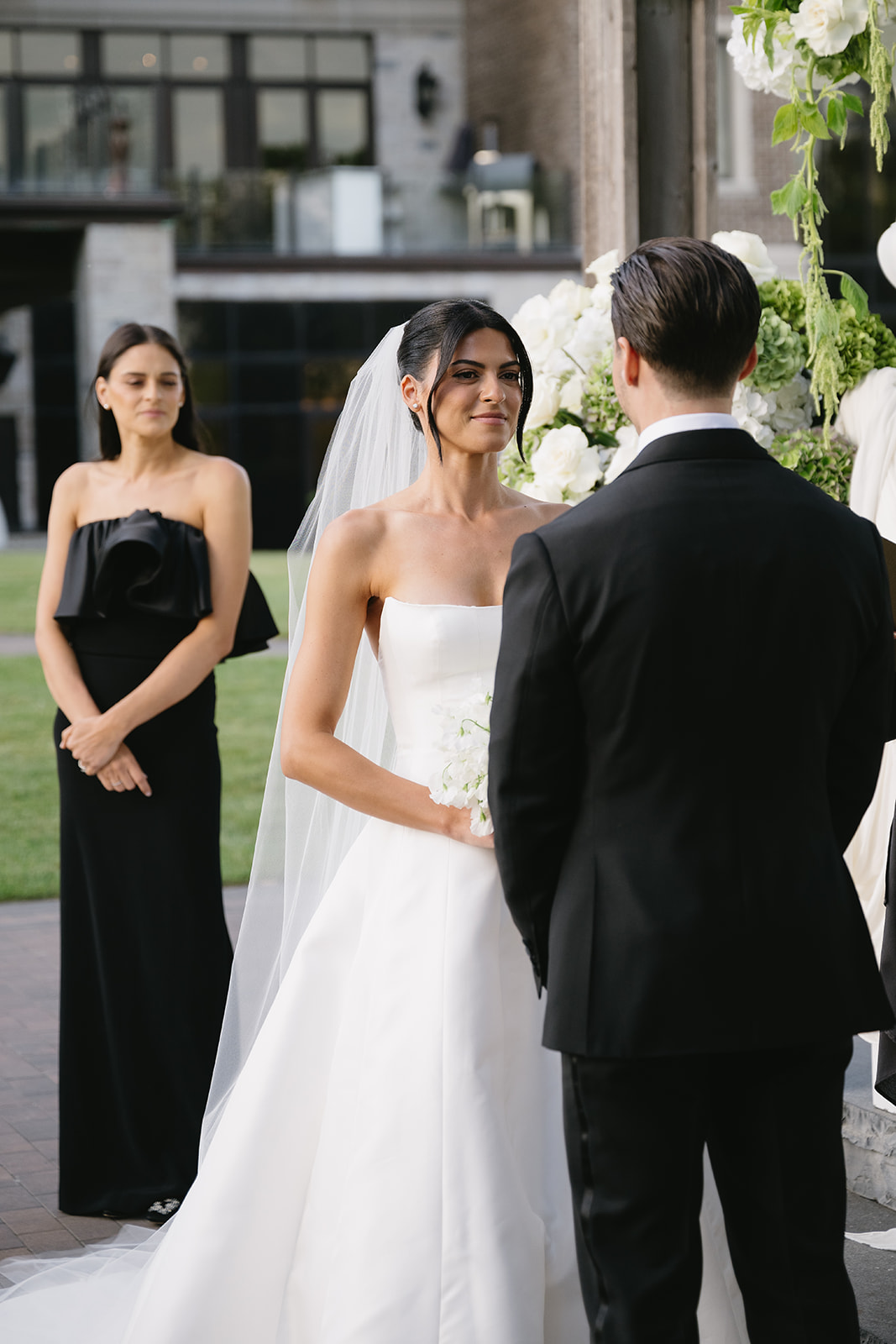 A bride in a white gown and veil stands facing a groom in a black suit during an outdoor wedding ceremony, with Erica, the bridesmaid in a black dress, standing nearby.