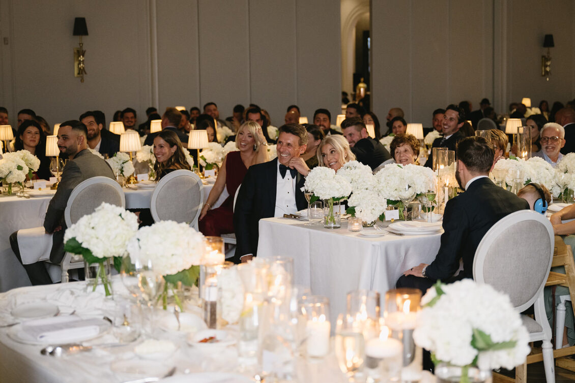 Guests in formal attire sit at tables decorated with white flowers and candles, smiling and watching something off-camera at an elegant indoor event or wedding reception—perhaps captivated by a heartfelt toast from Erica.