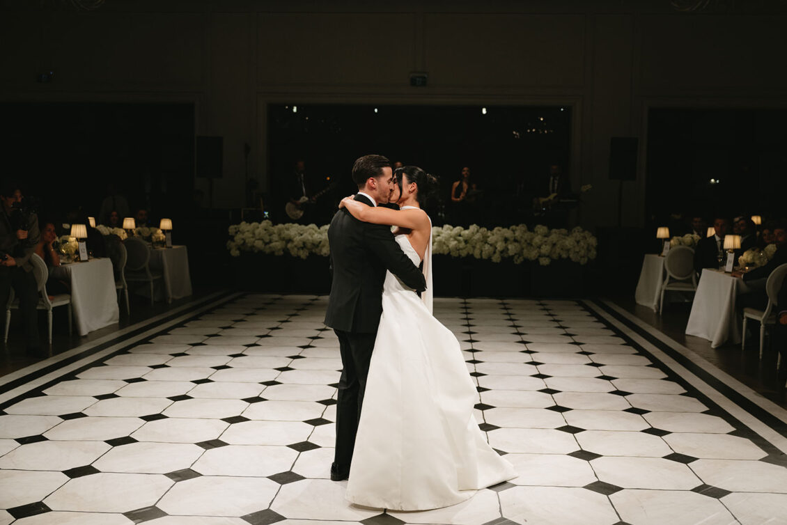 A bride and groom, Erica and her partner, share their first dance on a black-and-white tiled floor in a dimly lit, elegant venue with guests seated around the dance floor.
