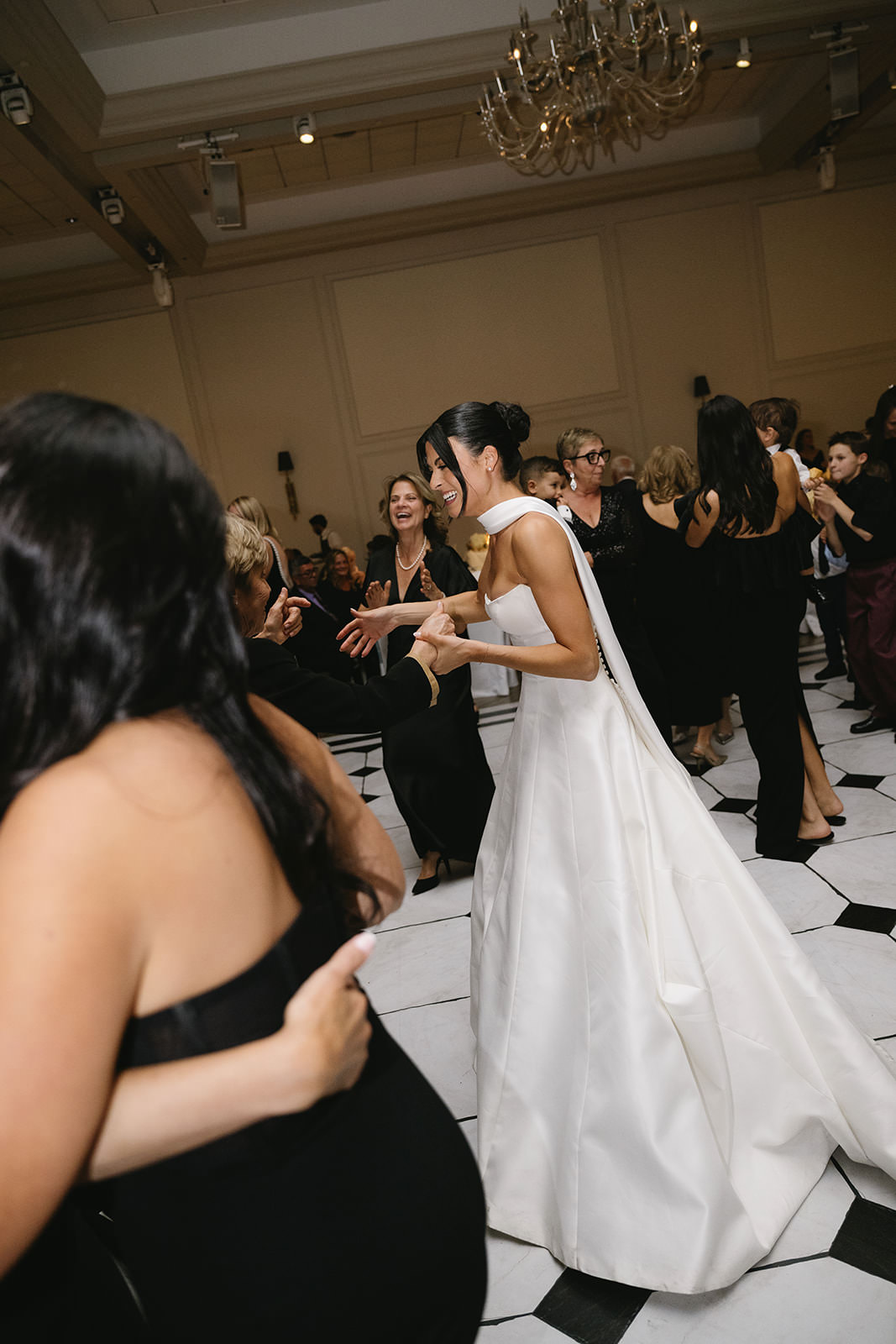 Erica, the radiant bride in a white dress, dances joyfully with guests on a black and white tiled floor at the wedding reception. People around her are smiling and enjoying the celebration.