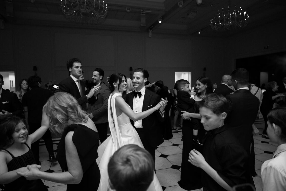 A bride and groom dance and smile at their wedding reception, surrounded by guests who are also dancing and celebrating in a formal, elegant venue—an unforgettable moment beautifully captured for Erica and her loved ones.