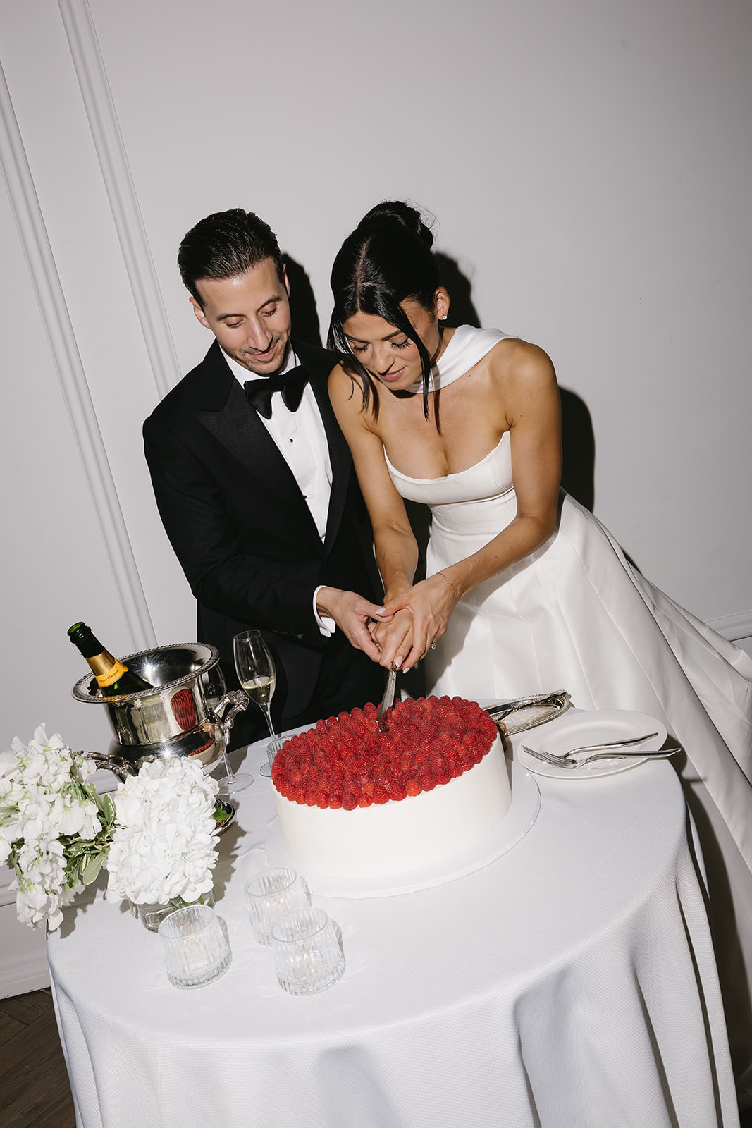A bride and groom, dressed formally, cut a white cake topped with berries together at a small round table with flowers, glasses, and champagne, as Erica looks on with joy.