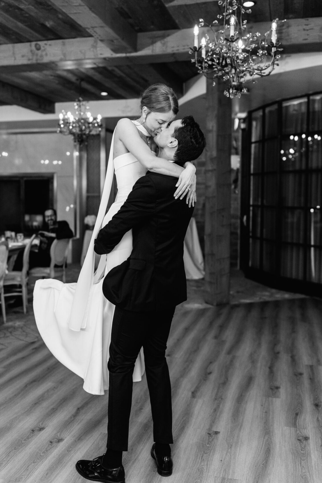 A man in a suit lifts a woman in a white dress as they embrace and smile joyfully in an elegant, chandelier-lit room with wooden floors, capturing the magic of an intimate Elora Mill wedding.
