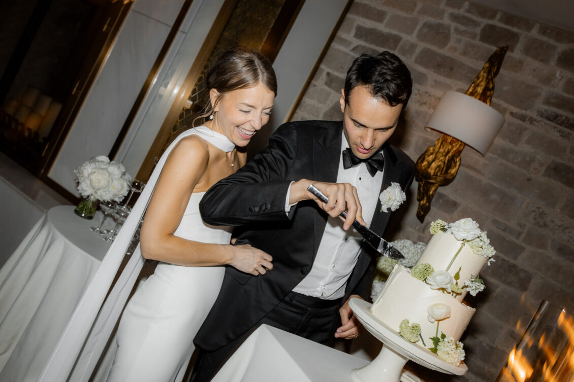 A bride and groom in formal attire smile and laugh as they cut a white, two-tier wedding cake decorated with flowers in an elegant indoor setting, capturing the joy of their intimate Elora Mill wedding.