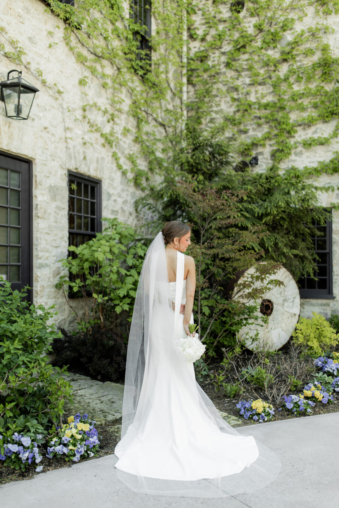 A bride in a white gown and veil stands outside a stone building with ivy, holding a bouquet and looking over her shoulder, surrounded by lush greenery and flowers—a perfect moment from an intimate Elora Mill wedding.