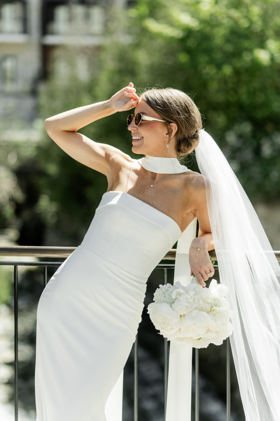 A bride in a strapless white wedding dress and veil leans on a railing, holding a bouquet of white flowers and wearing sunglasses, smiling outdoors on a sunny day at her intimate Elora Mill wedding.