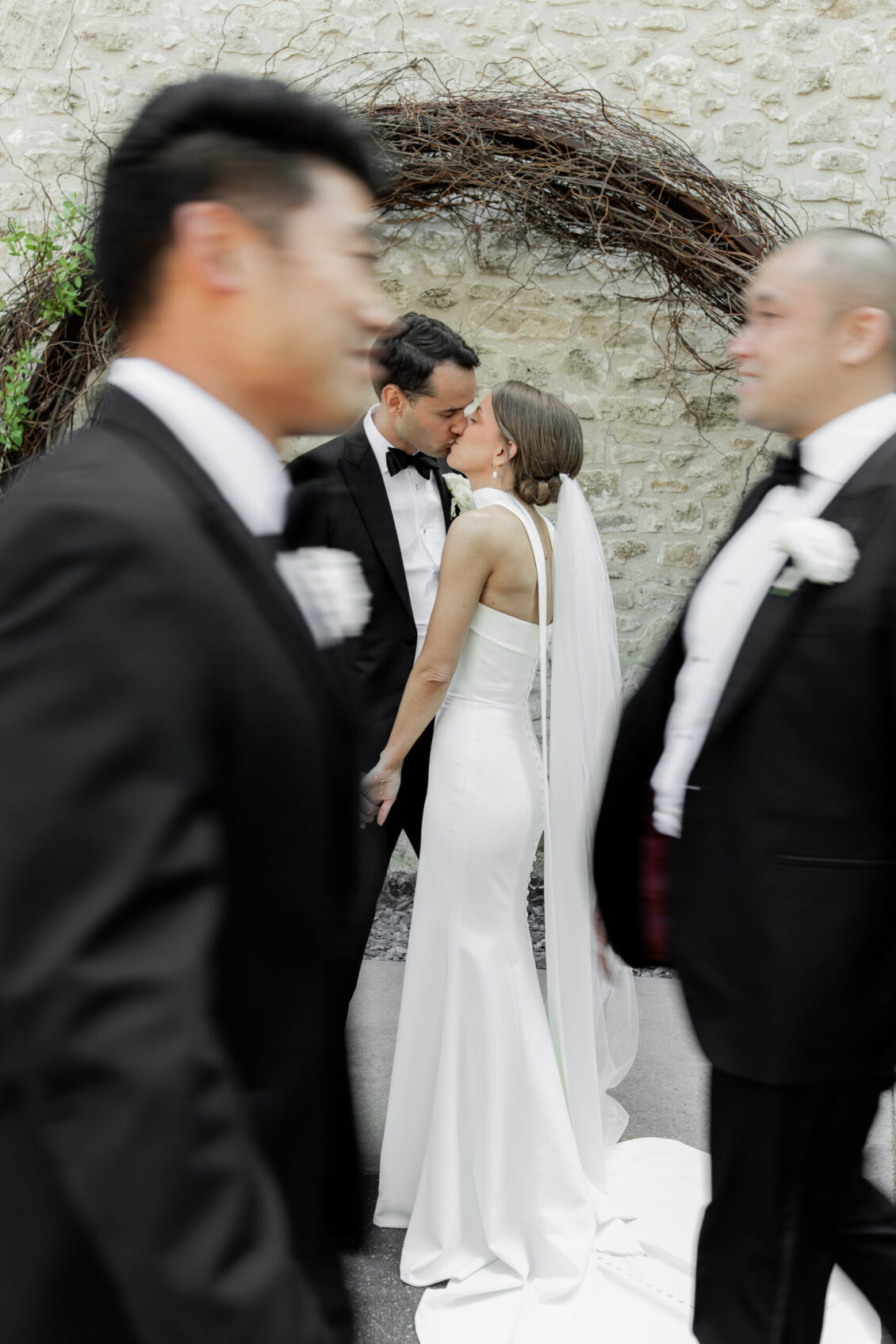 A bride and groom share an intimate kiss in front of a stone wall with a circular vine arch at their Elora Mill wedding, while two men in tuxedos walk past, slightly blurred in the foreground.