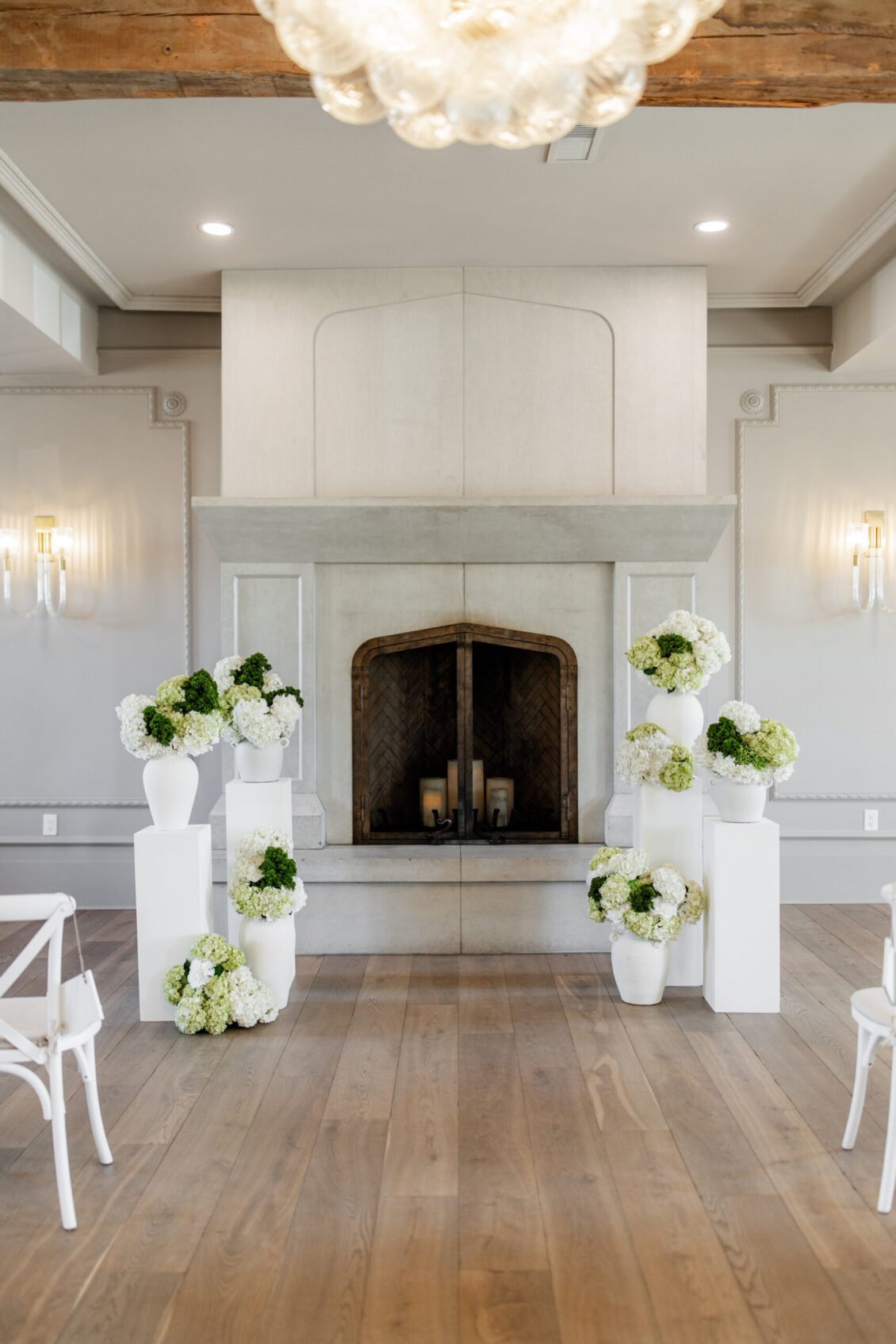 A bright room with a fireplace, white vases filled with white and green flowers on pedestals, wooden floor, white chairs, and soft lighting sets the perfect scene for an intimate Elora Mill wedding.
