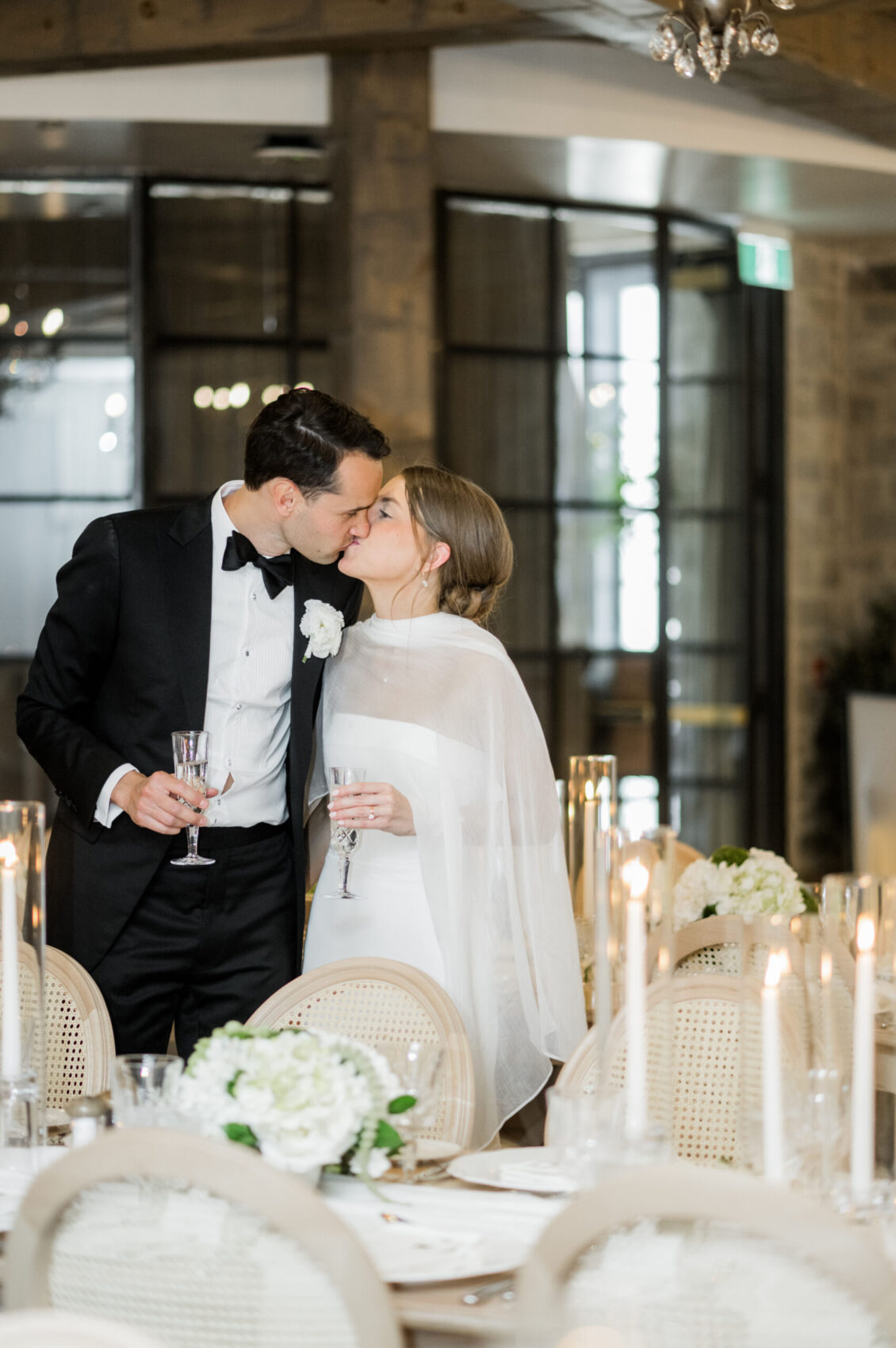 A bride and groom share a kiss while holding champagne glasses at a decorated wedding reception table with white flowers and candles, capturing the romance of their intimate Elora Mill wedding.