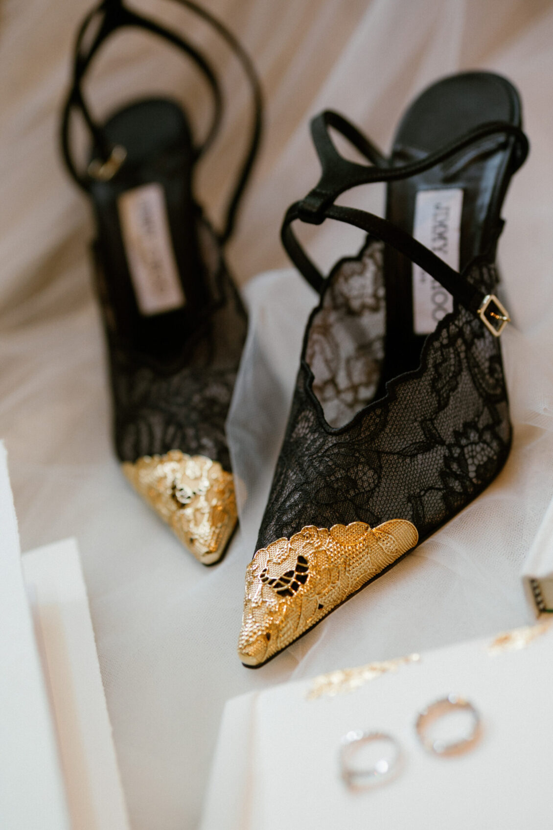Black lace high heels with delicate ankle straps, featuring pointed toes adorned with intricate gold embellishments—perfect for a romantic Renaissance Phuket wedding. The shoes are placed on a soft white fabric background near some jewelry, capturing an atmosphere of refined elegance.