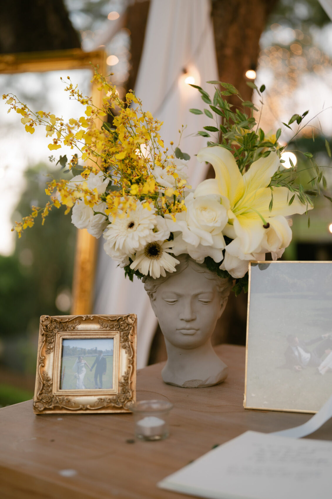 A floral arrangement in a head-shaped vase, next to a framed photo and another picture, sits on a wooden surface outdoors with trees and lights in the background, capturing the elegant ambiance of a Renaissance Phuket wedding.