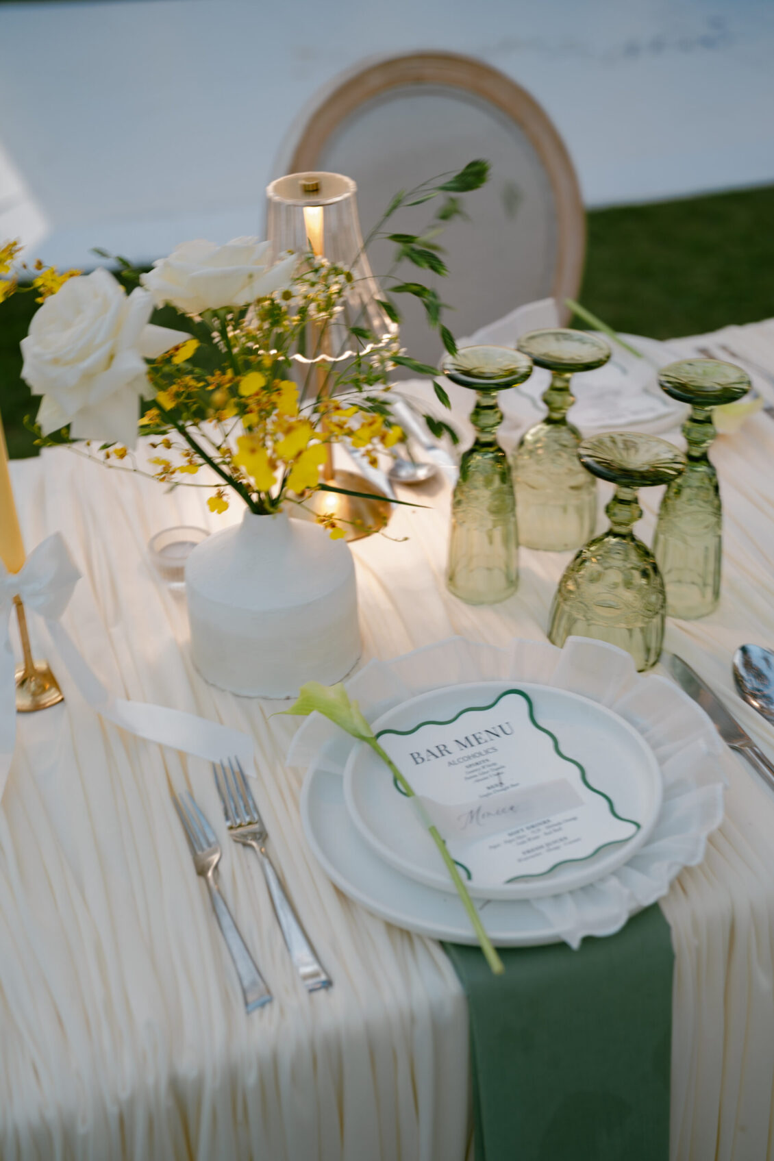 Elegant table setting for a Renaissance Phuket wedding, featuring white and yellow flowers in a vase, green glasses turned upside down, a white plate with a menu, silverware, and a candle on a cream-colored tablecloth.