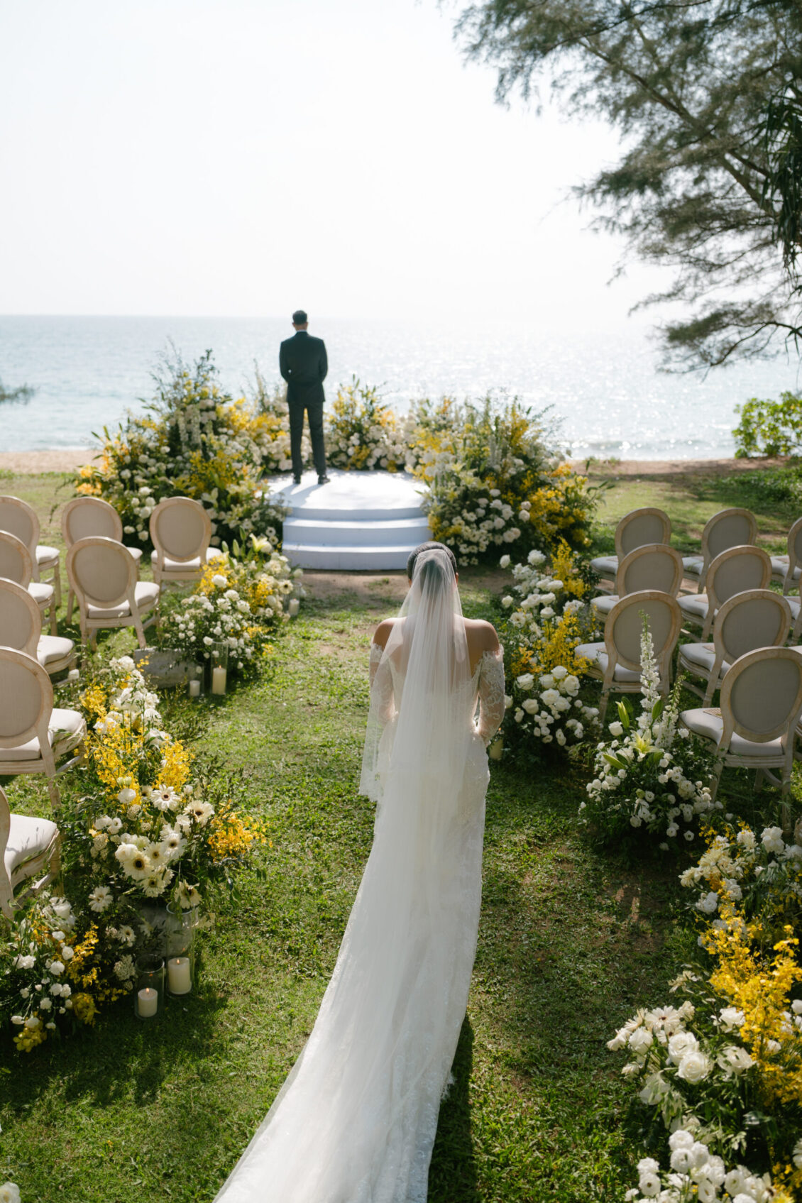 A bride in a white gown walks down an outdoor aisle lined with flowers toward a groom standing on a platform by the sea, framed by empty chairs on either side, capturing the romance and elegance of a Renaissance Phuket wedding.