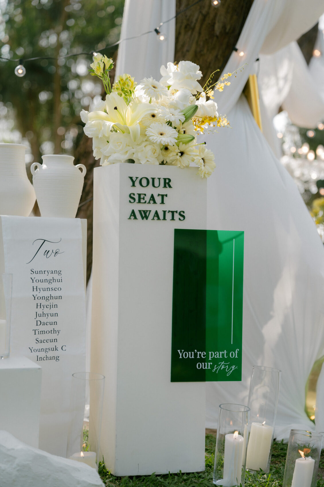 A white pedestal with green text reads “YOUR SEAT AWAITS.” It holds a bouquet of white and yellow flowers, evoking the fresh elegance of a Renaissance Phuket wedding. Nearby, glass vases, candles, and a guest name list are arranged, with draped white fabric and trees in the background.