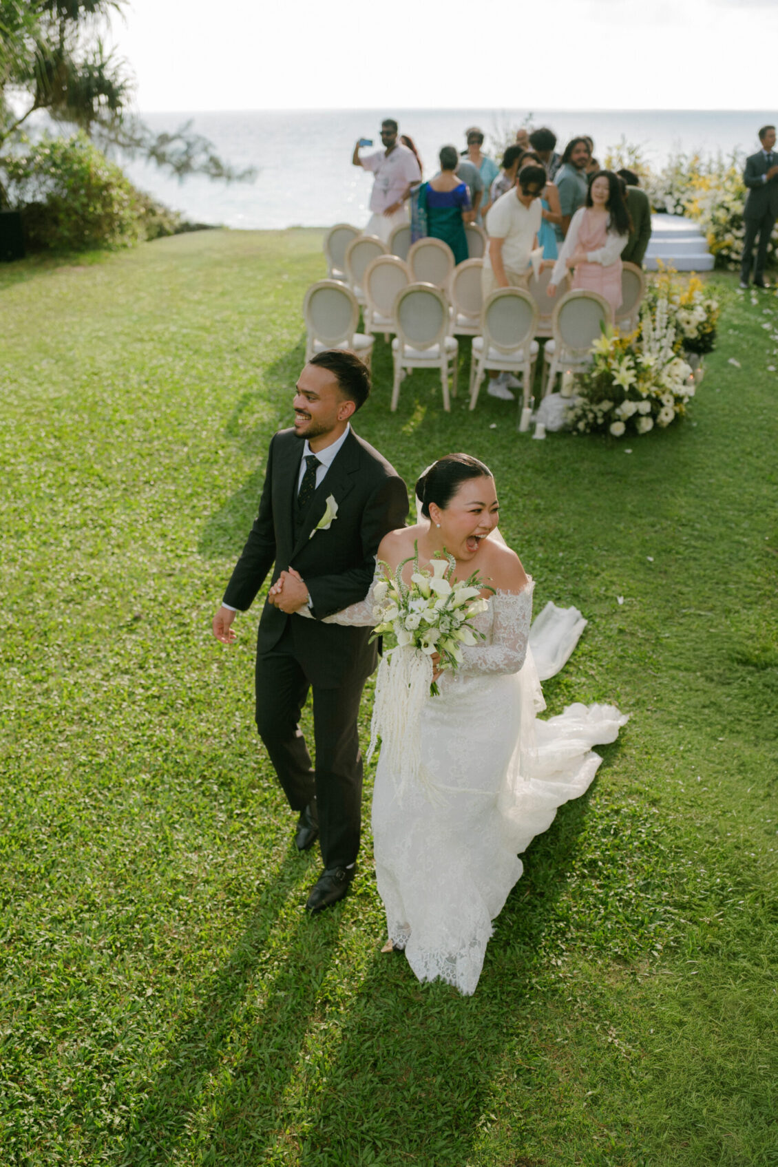 A newlywed couple walks hand in hand on grass, smiling joyfully, with wedding guests and white chairs in the background near the ocean at a picturesque Renaissance Phuket wedding.