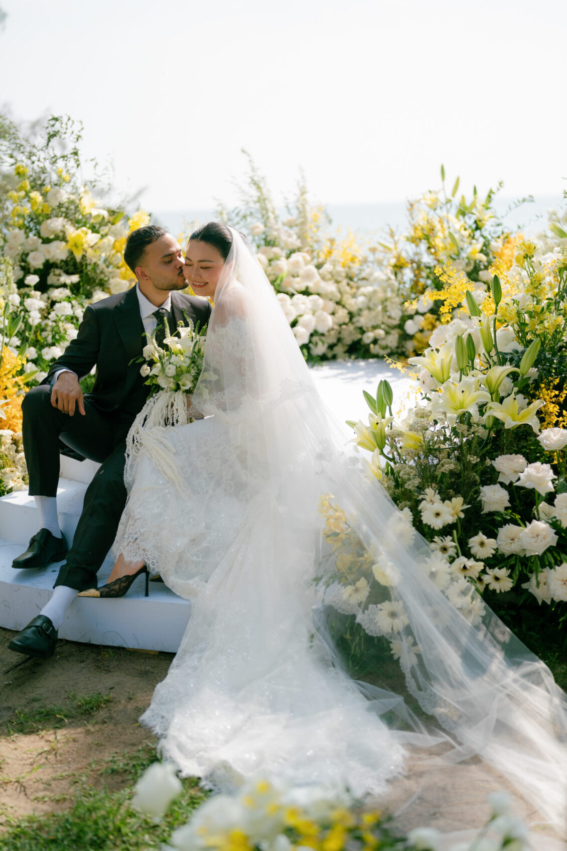 A bride and groom sit together, smiling and embracing amid lush white and yellow flowers at their Renaissance Phuket wedding ceremony. The bride wears a lace gown and veil, holding a bouquet.