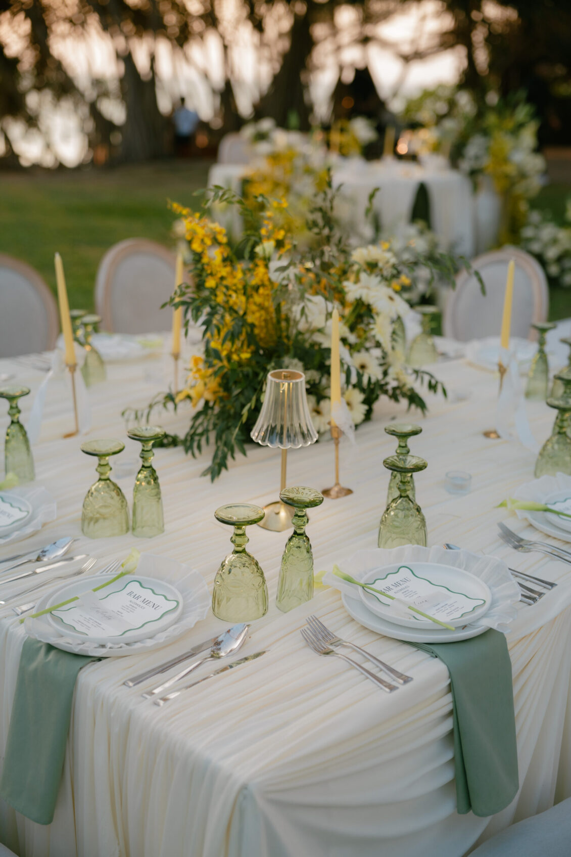 Outdoor table set for a Renaissance Phuket wedding event with green glassware, gold flatware, white plates with menus, green napkins, tall candles, and yellow-and-white floral centerpieces. Trees and blurred greenery are in the background.