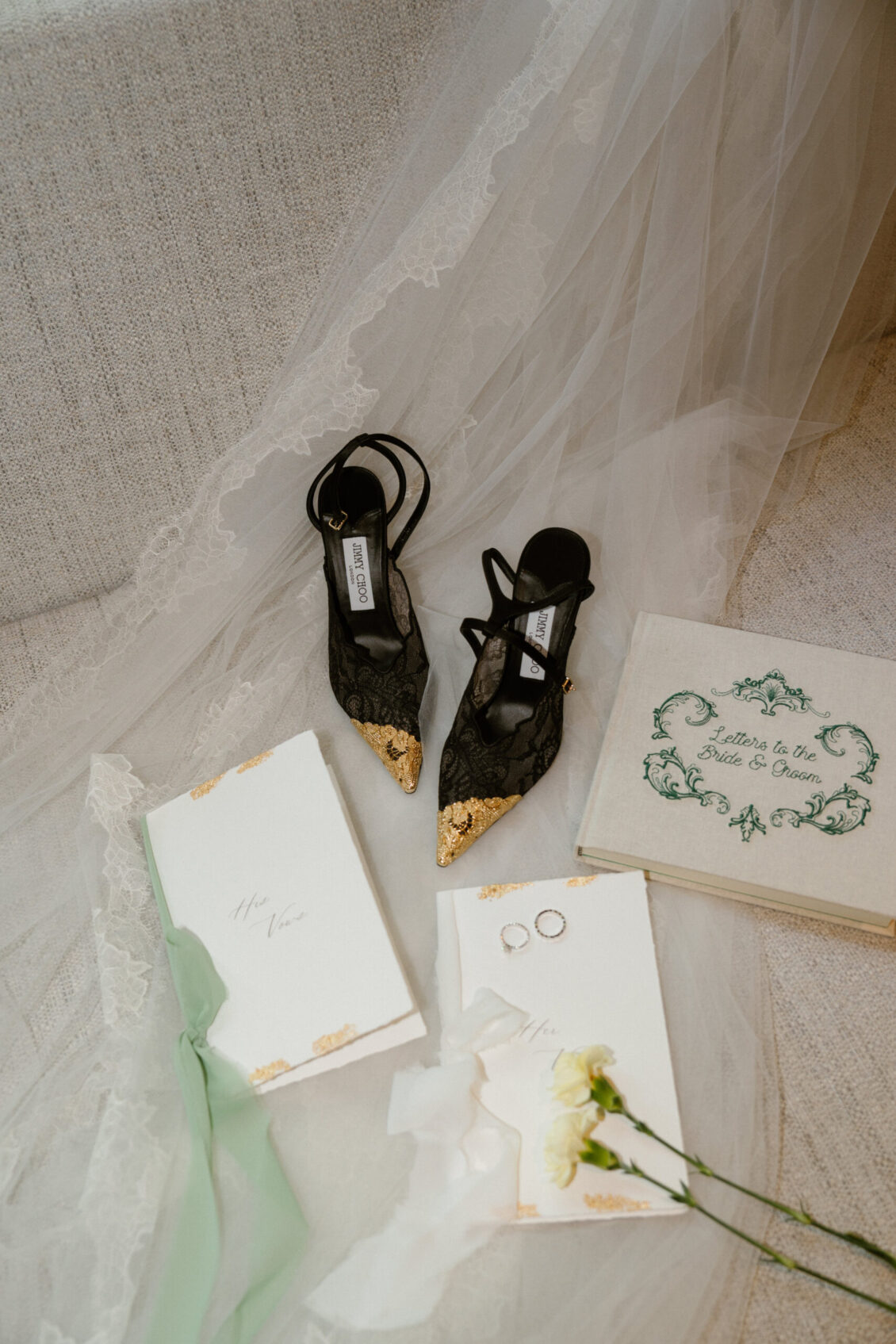 Black pointed lace heels, two gold-trimmed invitations with green ribbon, two rings, a white carnation, an ornate book, and a lace veil arranged on a beige fabric surface evoke the refined elegance of a Renaissance Phuket wedding.