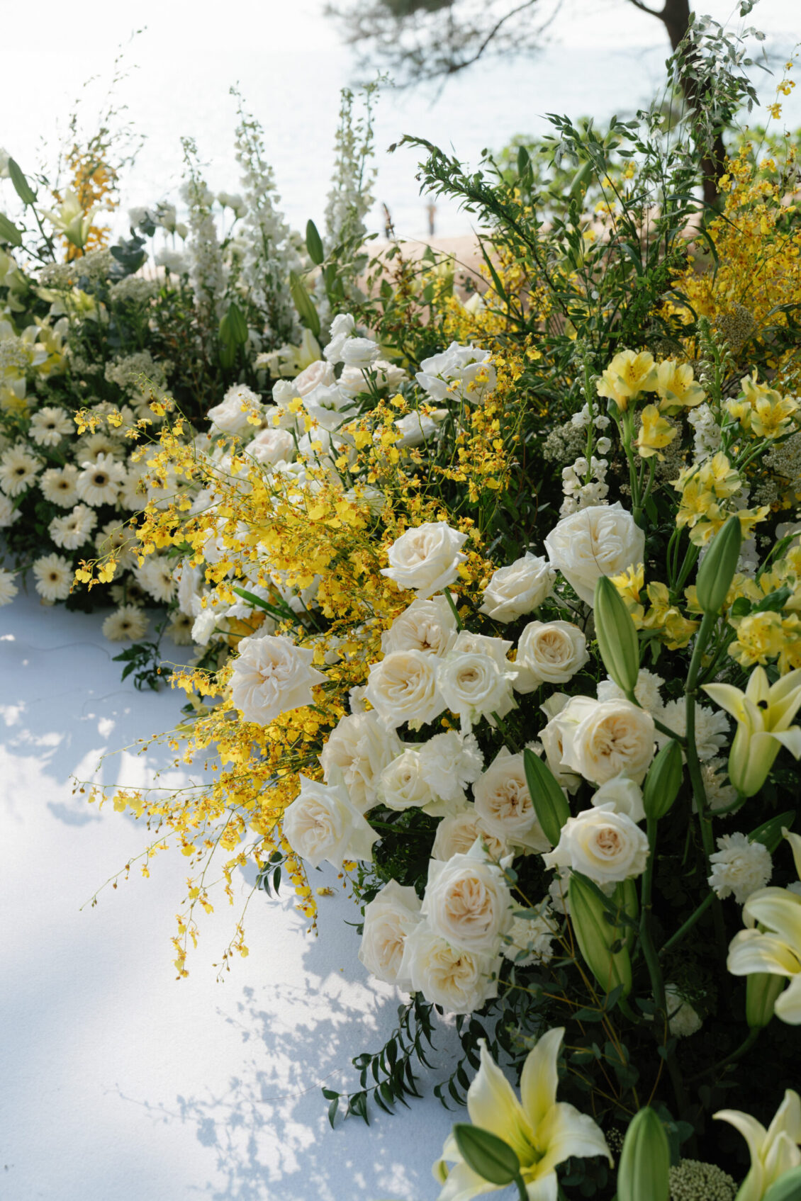 A lush arrangement of white and yellow flowers, including roses, lilies, and daisies, set outdoors on a sunny day with greenery and trees in the background—perfect for a picturesque Renaissance Phuket wedding.
