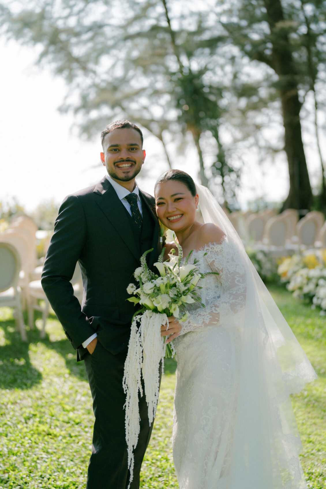 A bride and groom stand together outdoors at their Renaissance Phuket wedding, smiling at the camera. The bride holds a bouquet of white flowers and wears a white lace gown, while the groom is dressed in a dark suit. Trees and chairs are visible in the background.