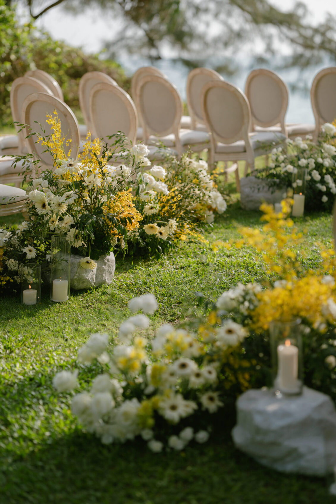Rows of elegant white chairs and floral arrangements with white and yellow flowers and candles are set on a green lawn, creating a serene Renaissance Phuket wedding setup. Trees and blurred water are visible in the background, adding to the tranquil ambiance of this outdoor event.