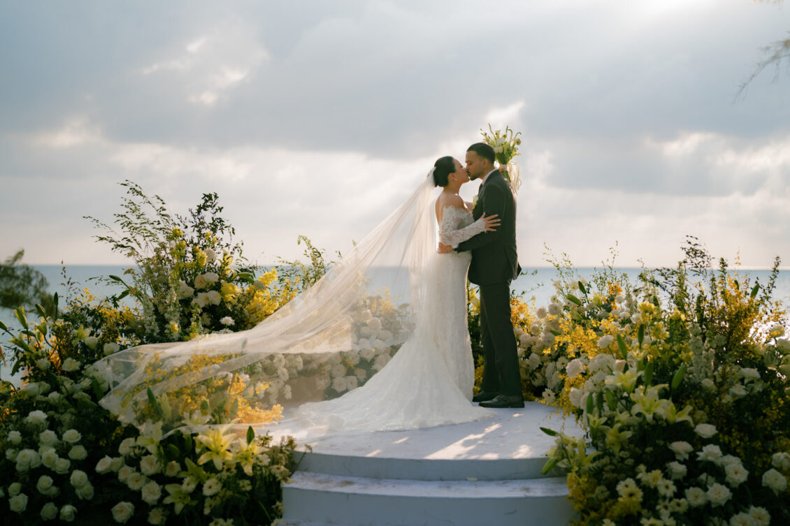 A bride and groom kiss on an outdoor platform at their Renaissance Phuket wedding, surrounded by lush yellow and white flowers, with the ocean and a cloudy sky in the background. The bride’s veil flows behind her.