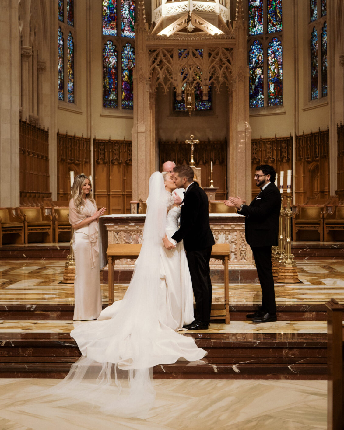 A bride and groom kiss at the altar during their church wedding ceremony, while two attendants stand nearby and one person applauds. Stained glass windows and ornate architecture are visible in the background, capturing the elegance of Ariana and Julian Borghese’s wedding.