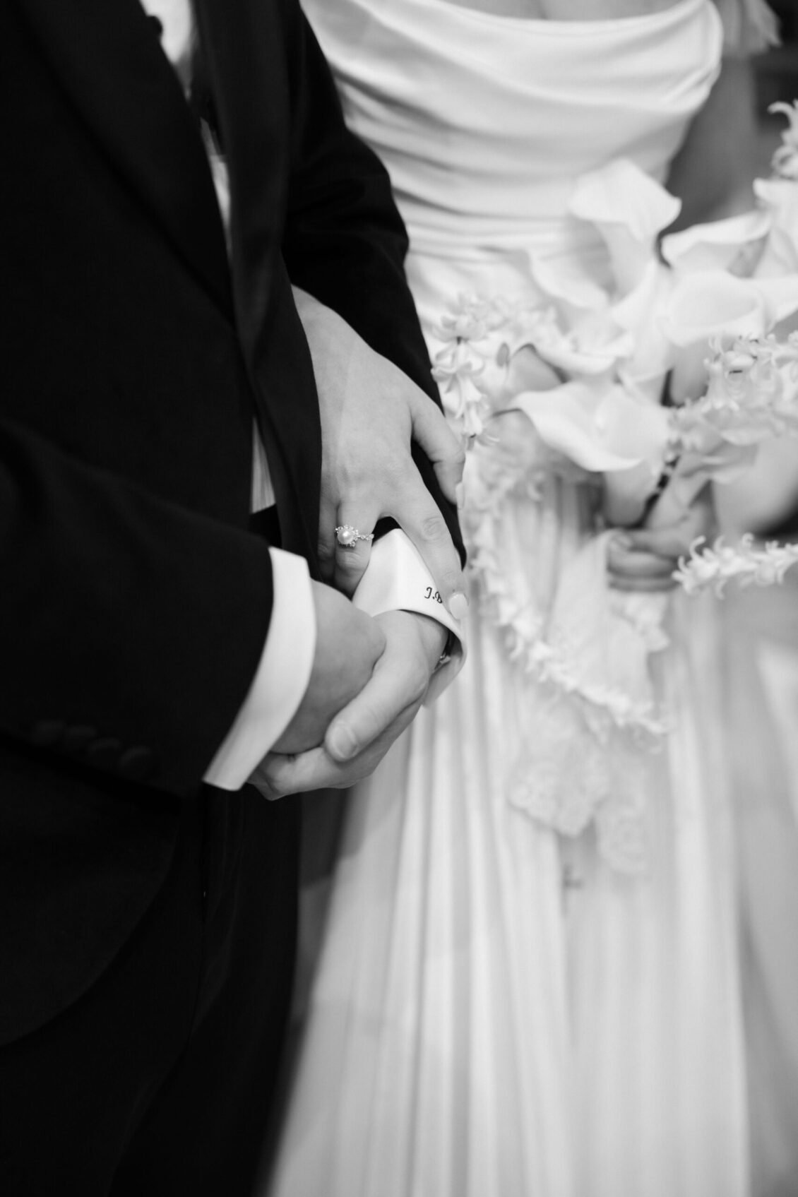 A bride and groom stand close together at the Ariana and Julian Borghese wedding. The bride holds a bouquet of flowers and gently holds the groom’s hand, showing a wedding ring. Both wear formal attire. The image is black and white.