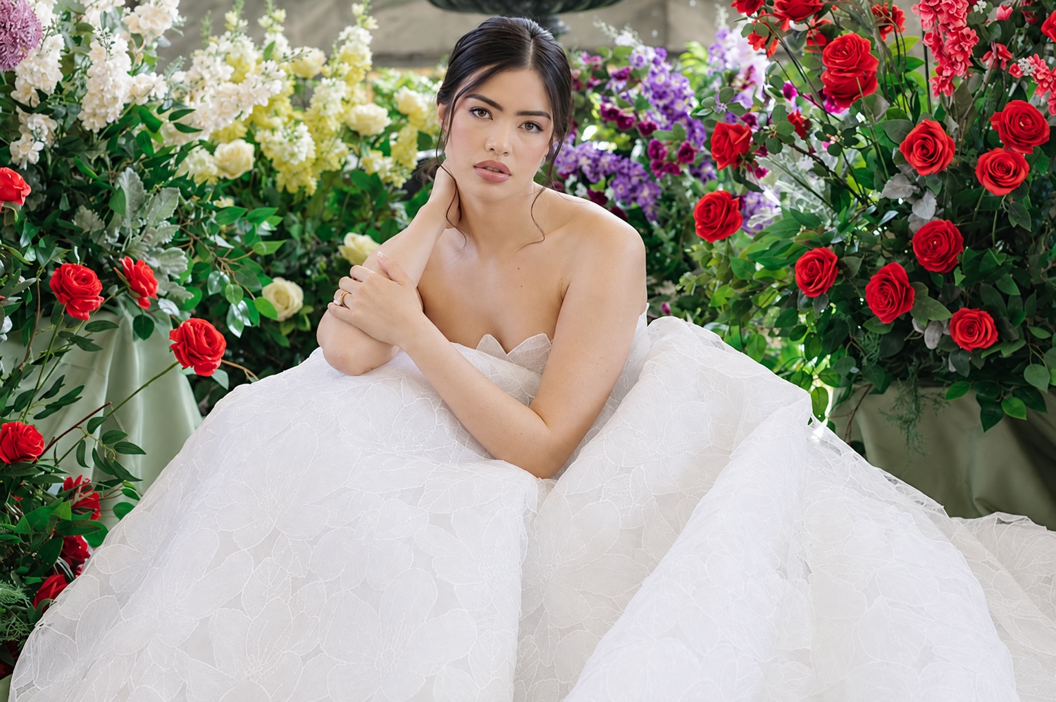 A woman in a strapless white gown sits among vibrant red, yellow, and purple flowers, gazing thoughtfully at the camera in this enchanting Casa Loma wedding editorial.