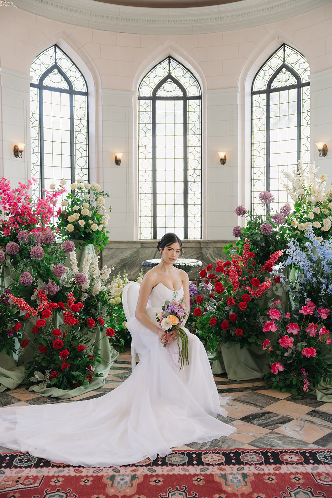 A bride in a white gown sits on a chair, holding a bouquet, surrounded by vibrant red, pink, and purple flowers in front of large arched windows in an elegant, sunlit room—a scene reminiscent of a Casa Loma wedding editorial.