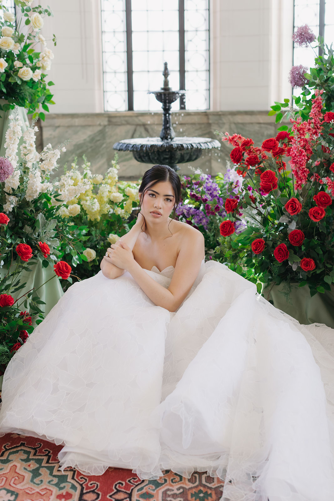A woman in a white wedding gown sits surrounded by vibrant red, purple, and yellow flowers in front of a window and a black fountain, looking thoughtfully at the camera in this elegant Casa Loma wedding editorial.
