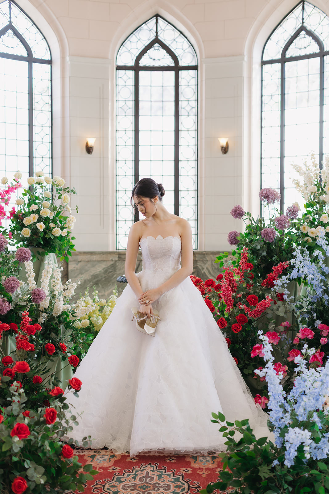 A bride in a white gown stands among colorful flowers, holding her shoes and looking down, with tall arched windows and soft natural light in the background—capturing the romantic essence of a Casa Loma wedding editorial.