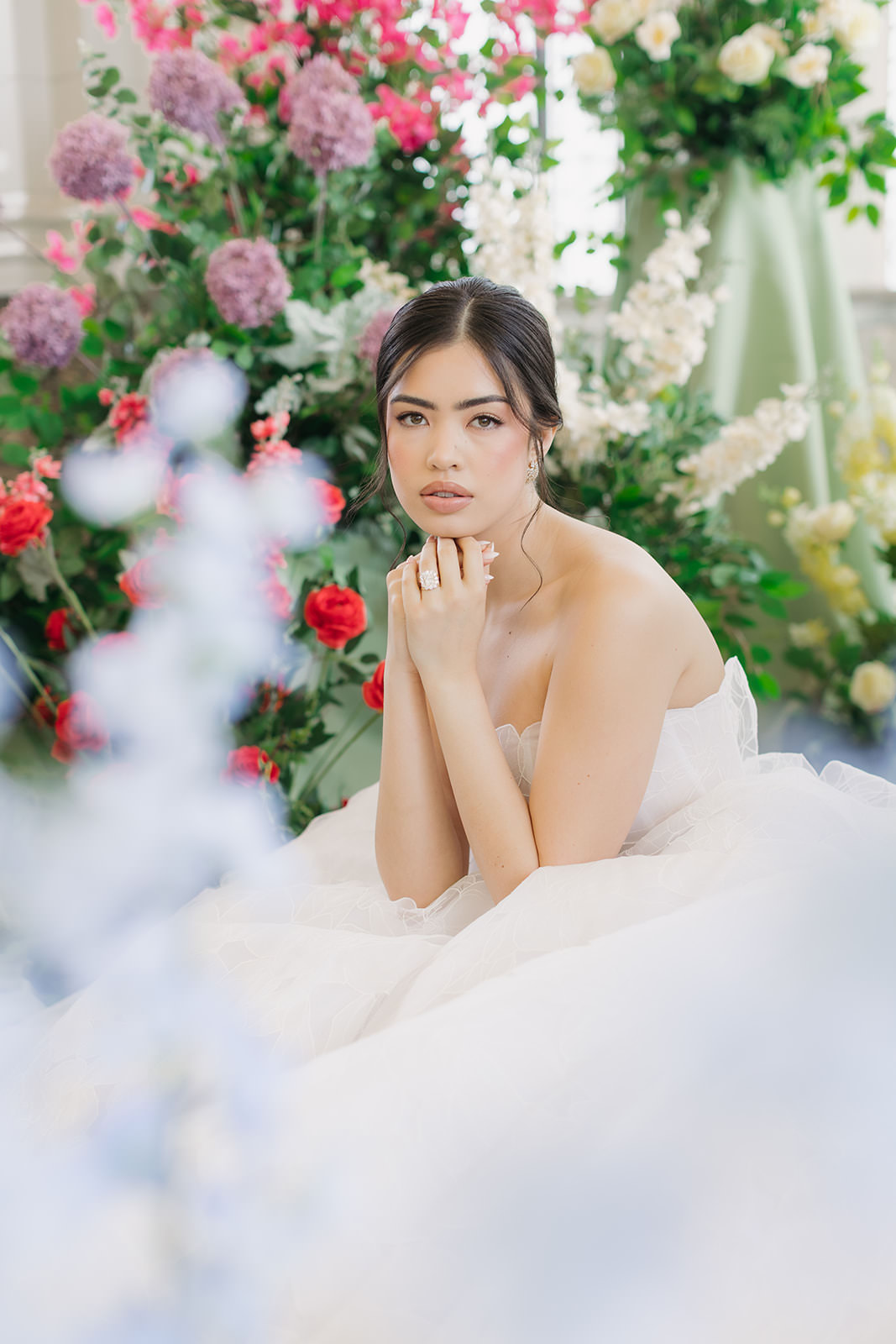 A woman in a white strapless gown sits surrounded by colorful flowers, resting her chin on her hands and gazing softly at the camera in a romantic Casa Loma wedding editorial.