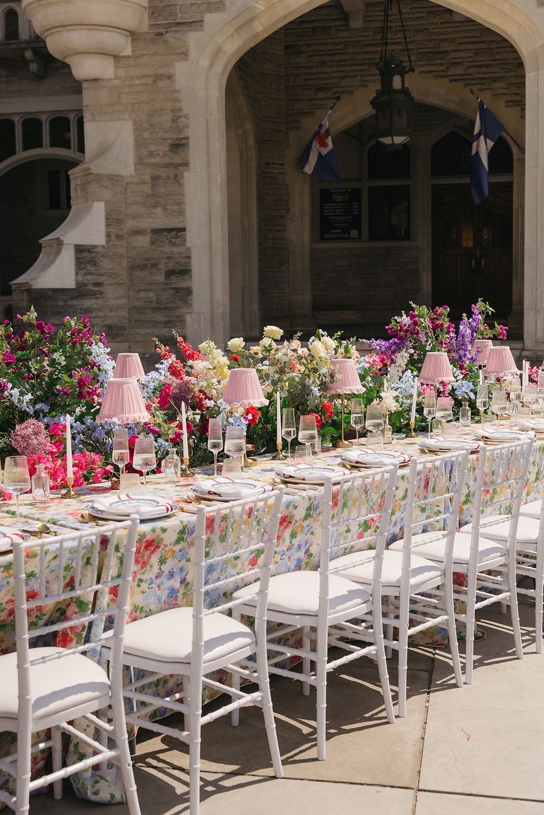 A long, elegant outdoor table is set with a floral tablecloth, pink lamps, glassware, and surrounded by white chairs. Colorful flowers line the center, with a stone building and arches in the background, capturing the romantic charm of a Casa Loma wedding editorial.