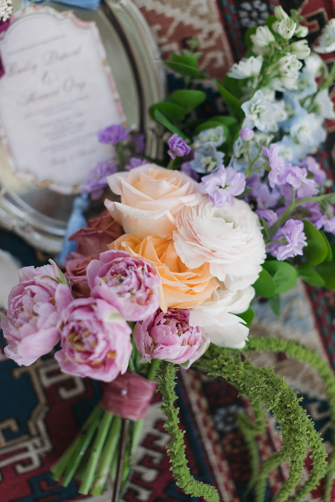 A bouquet of pink, peach, and white flowers with greenery lies on a patterned rug next to a silver tray holding a partially blurred invitation, capturing the romantic elegance of a Casa Loma wedding editorial.