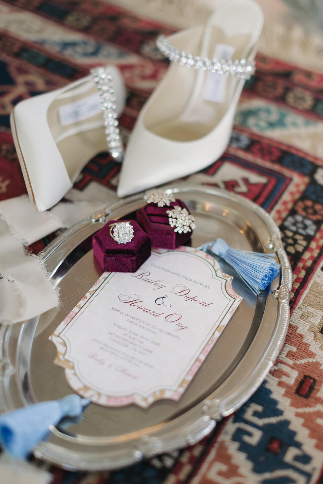 A silver tray holds a wedding invitation, two red ring boxes with diamond rings, and blue tassels, capturing the refined elegance of a Casa Loma wedding editorial. White high-heeled shoes with rhinestone straps rest in the background on a patterned rug.