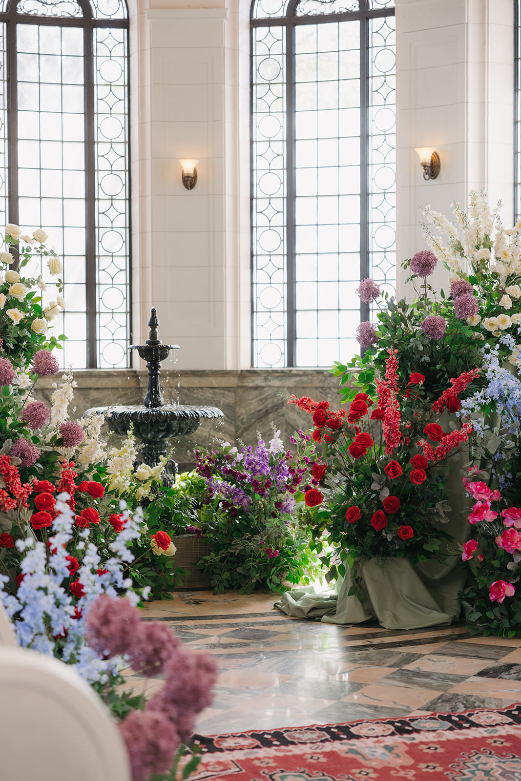 A grand indoor space with tall windows, a black fountain, and abundant floral arrangements in red, purple, pink, white, and blue—displayed on marble floors and near a patterned rug—evokes the timeless romance of a Casa Loma wedding editorial.