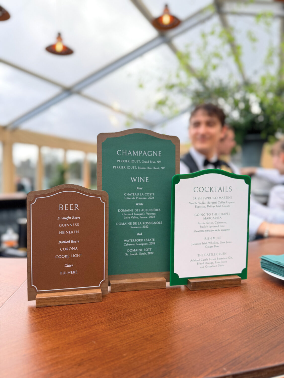 Three menu cards on a table list beer, champagne, wine, and cocktail options at an outdoor event. A bartender stands in the blurred background under a clear tent with greenery, capturing the elegant ambiance of an Irish castle wedding at Ashford Castle.