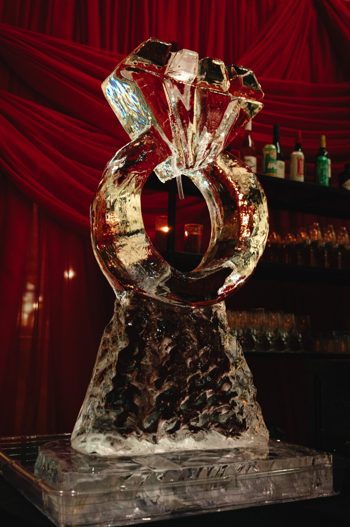 A carved diamond-ring ice sculpture glows gold and red in front of gathered red fabric and a dim bar.