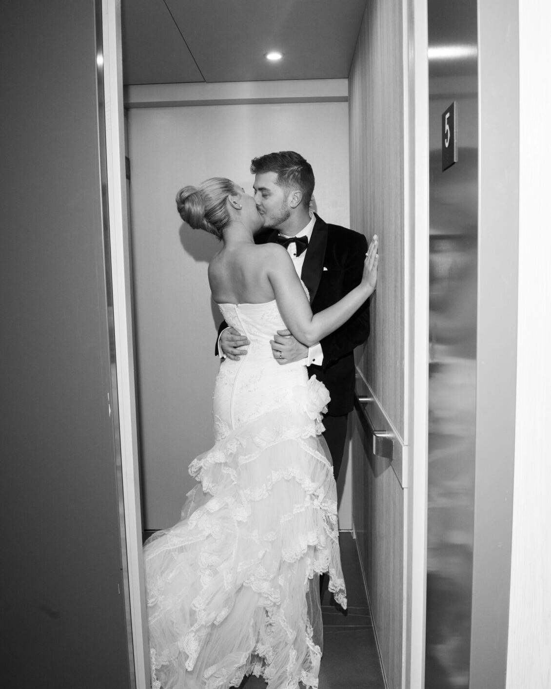A bride in a wedding dress and a groom in a tuxedo share a kiss inside an open elevator, capturing a romantic moment from the Ariana and Julian Borghese wedding.