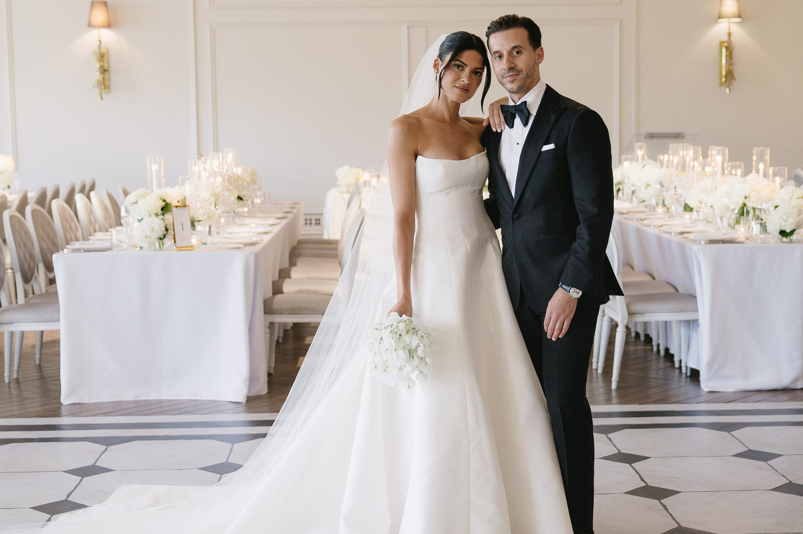 Bride Erica in a white gown and groom in a black tuxedo pose together in an elegant, white-themed wedding reception room decorated with flowers and candles.