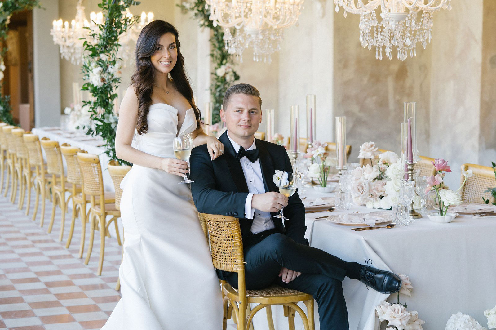 Bride in a white gown stands beside a groom in a black tuxedo, seated at an elegant, flower-adorned reception table with chandeliers overhead. Both hold wine glasses and smile at the camera, capturing the romance of a Tuscany villa wedding.