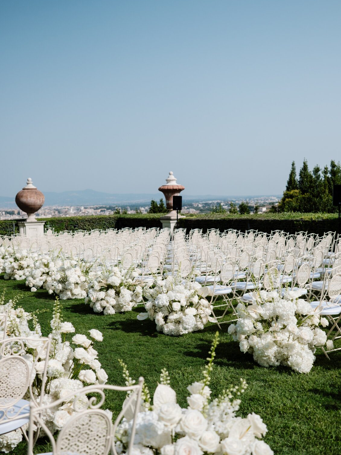 Rows of white chairs and lush white floral arrangements are set up outdoors on a green lawn for a Nigerian wedding in Rome, with a scenic city view and clear blue sky in the background.