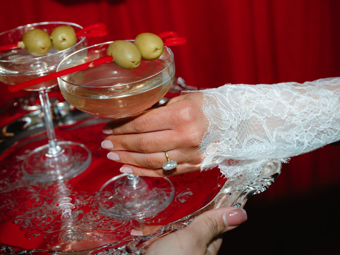 A hand in a lace sleeve with a ring holds a silver tray with two martini glasses, each garnished with two green olives, against a red background—a sophisticated scene reminiscent of a black-tie wedding at Hotel X Toronto.