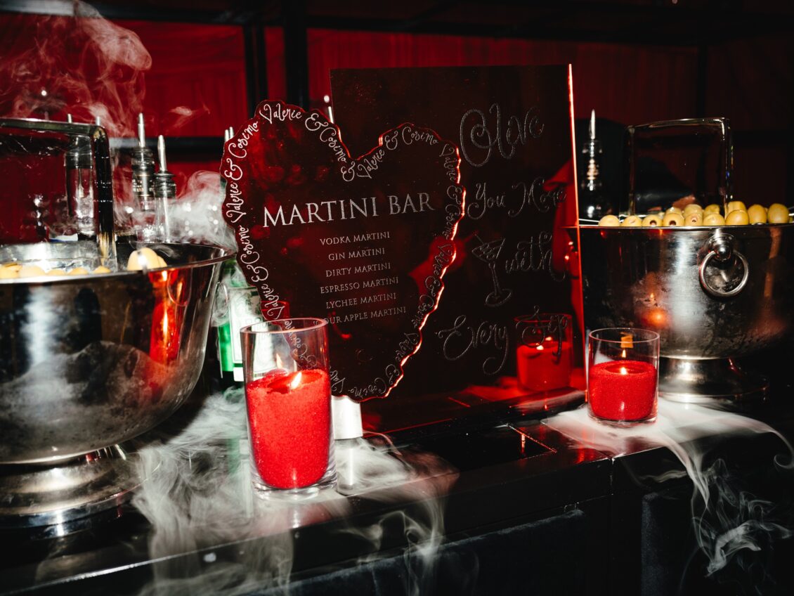 A black Martini Bar menu stands among bottles, ice buckets, olives, red drinks, candles, and curling white vapor.