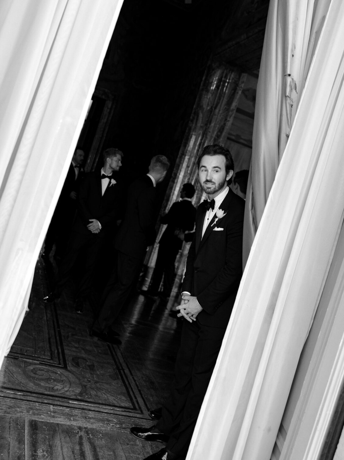 Groom stands between billowing curtains with attendants behind him.